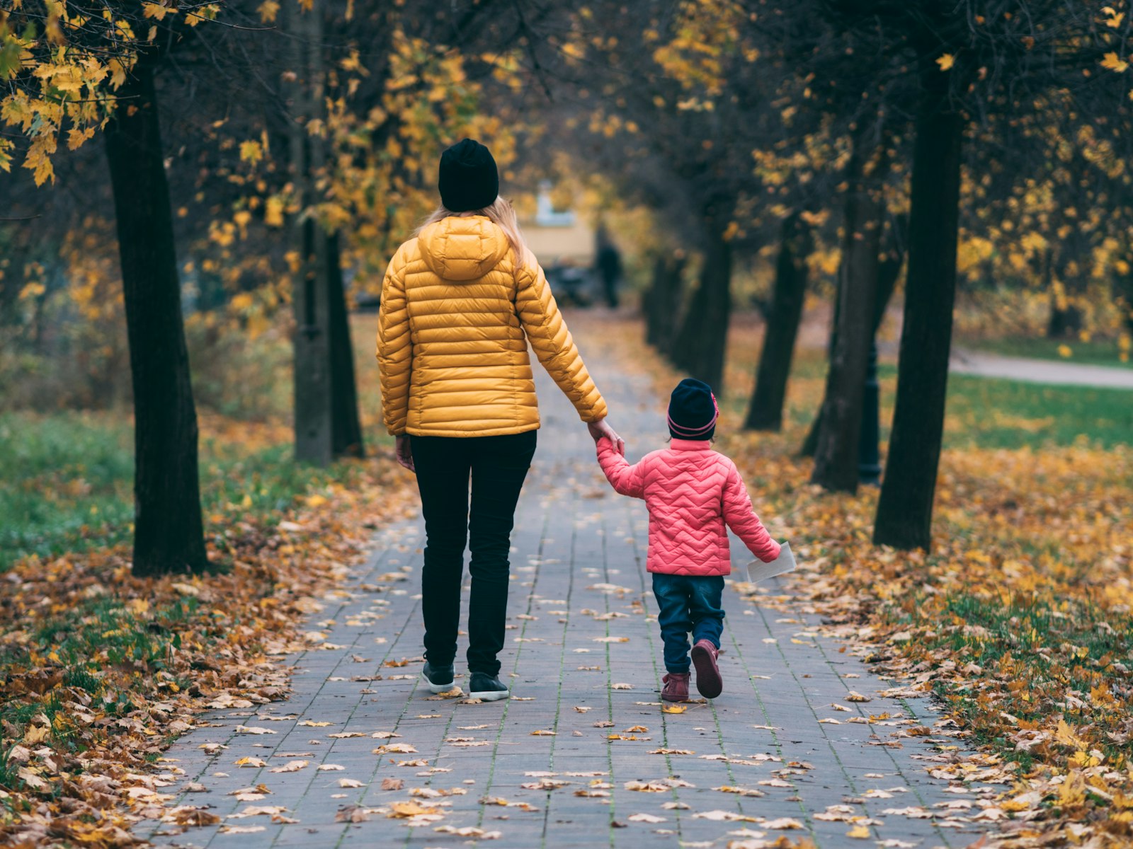 A parent and young child walking hand in hand down a leaf-covered path in autumn